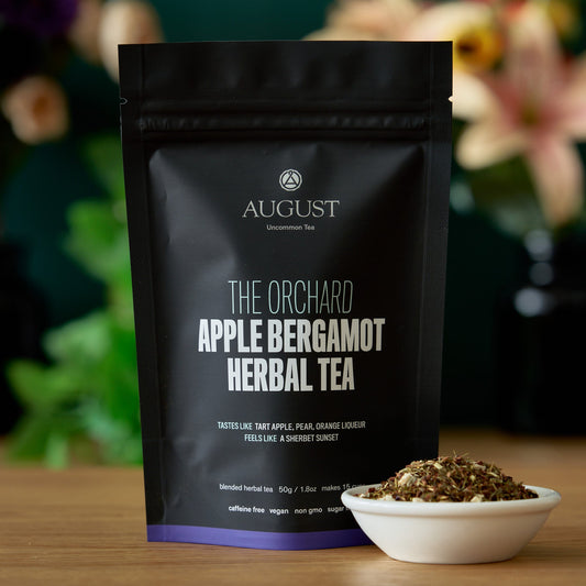 The Orchard Apple Bergamot Herbal Tea packaging with tea leaves, featuring tart apple and pear flavors, caffeine free.
