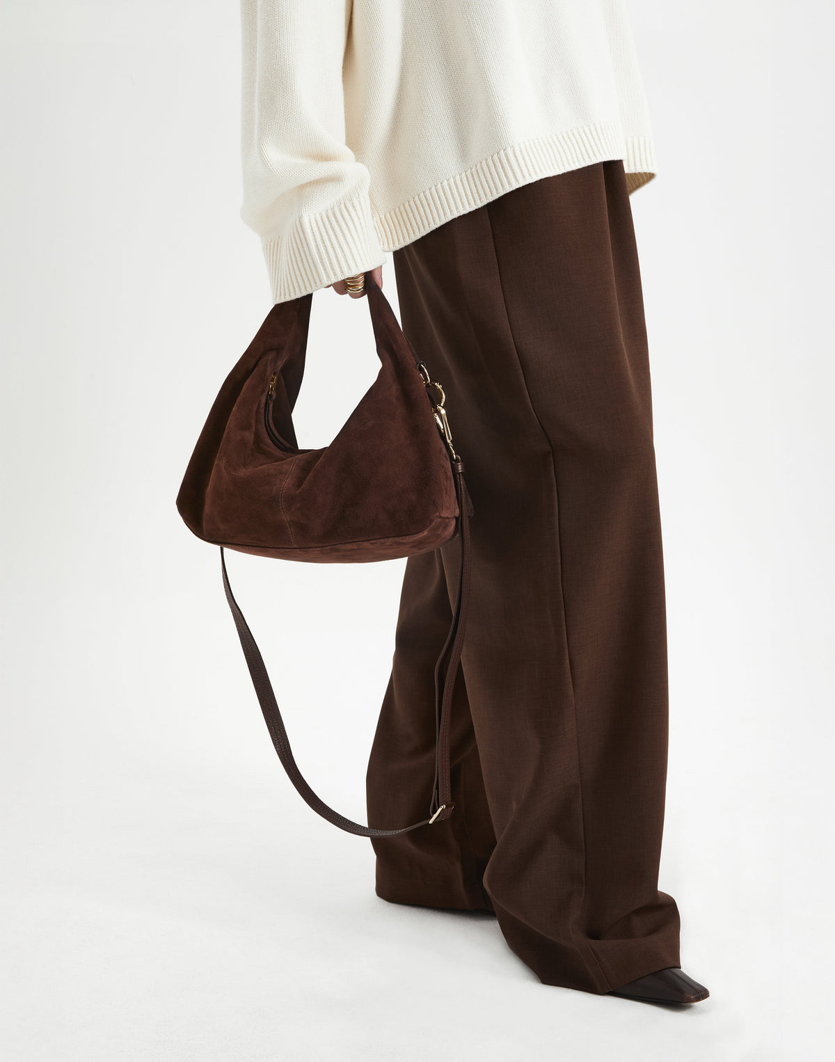 Woman holding a Mini Everyday Shoulder Bag made of soft brown suede, styled with a cozy white sweater and brown pants.