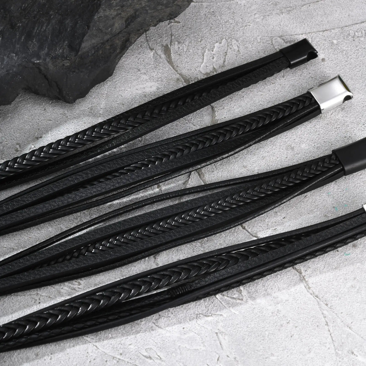Black mens braided leather bracelet featuring multi-layer straps and magnetic clasps on a grey stone surface.